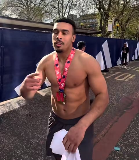 Man with muscular build wearing red medal lanyard standing shirtless outdoors at what appears to be an event or competition venue