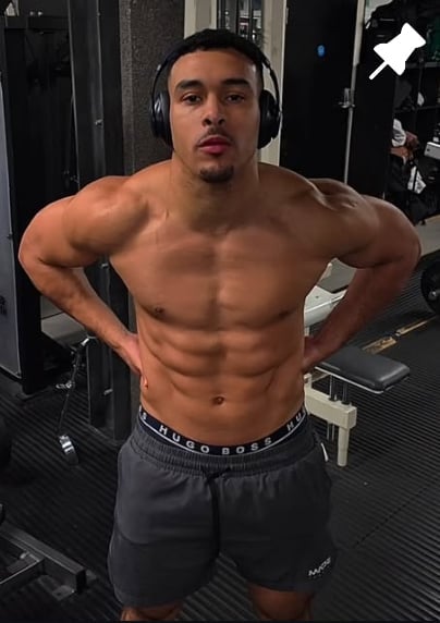 Shirtless muscular man wearing headphones and dark shorts posing in a gym with hands on hips